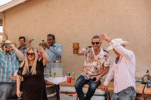 People laughing and enjoying drinks at a casual outdoor social gathering against a beige wall.