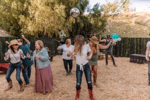 Group of people line dancing outdoors in a rustic setting with trees and decorative string lights.