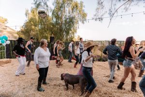 Line dancers in western attire moving in sync at an outdoor gathering with autumn trees.