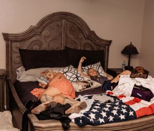 Multiple people sleeping on a large bed with an ornate headboard and American flag blanket.