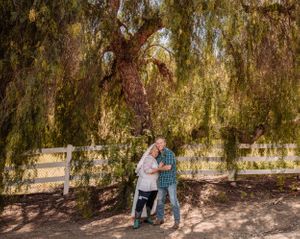 A couple shares a tender moment under a large weeping willow tree beside a wooden fence.