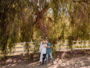 Two people embrace while standing beneath the drooping branches of a willow tree.