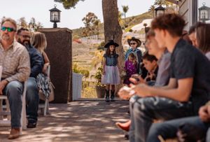Outdoor gathering with guests seated on benches under tree with lantern decorations.