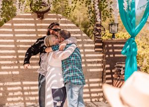 A couple embraces in a rustic outdoor setting with wooden beams casting shadows.