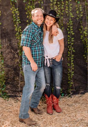 Playful couple in western-inspired outfits posing against vine wall backdrop.