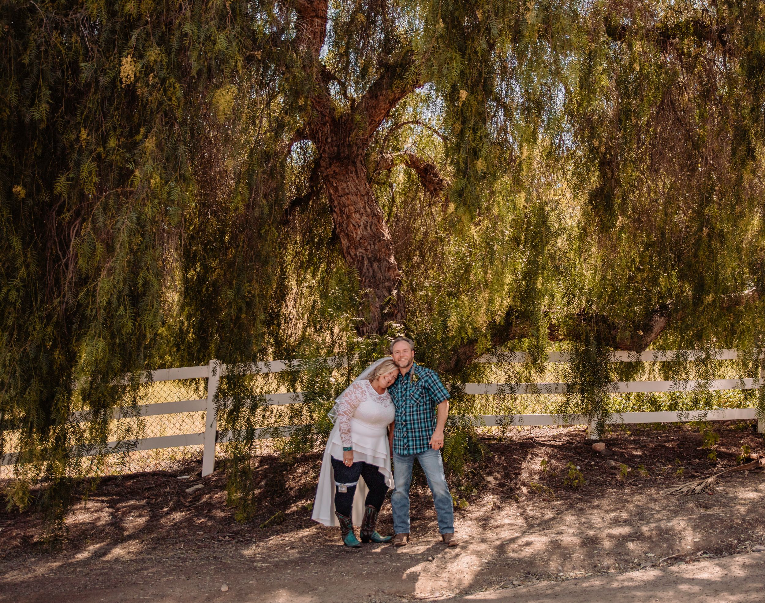 Two people pose together under the cascading branches of a willow tree near a white fence.