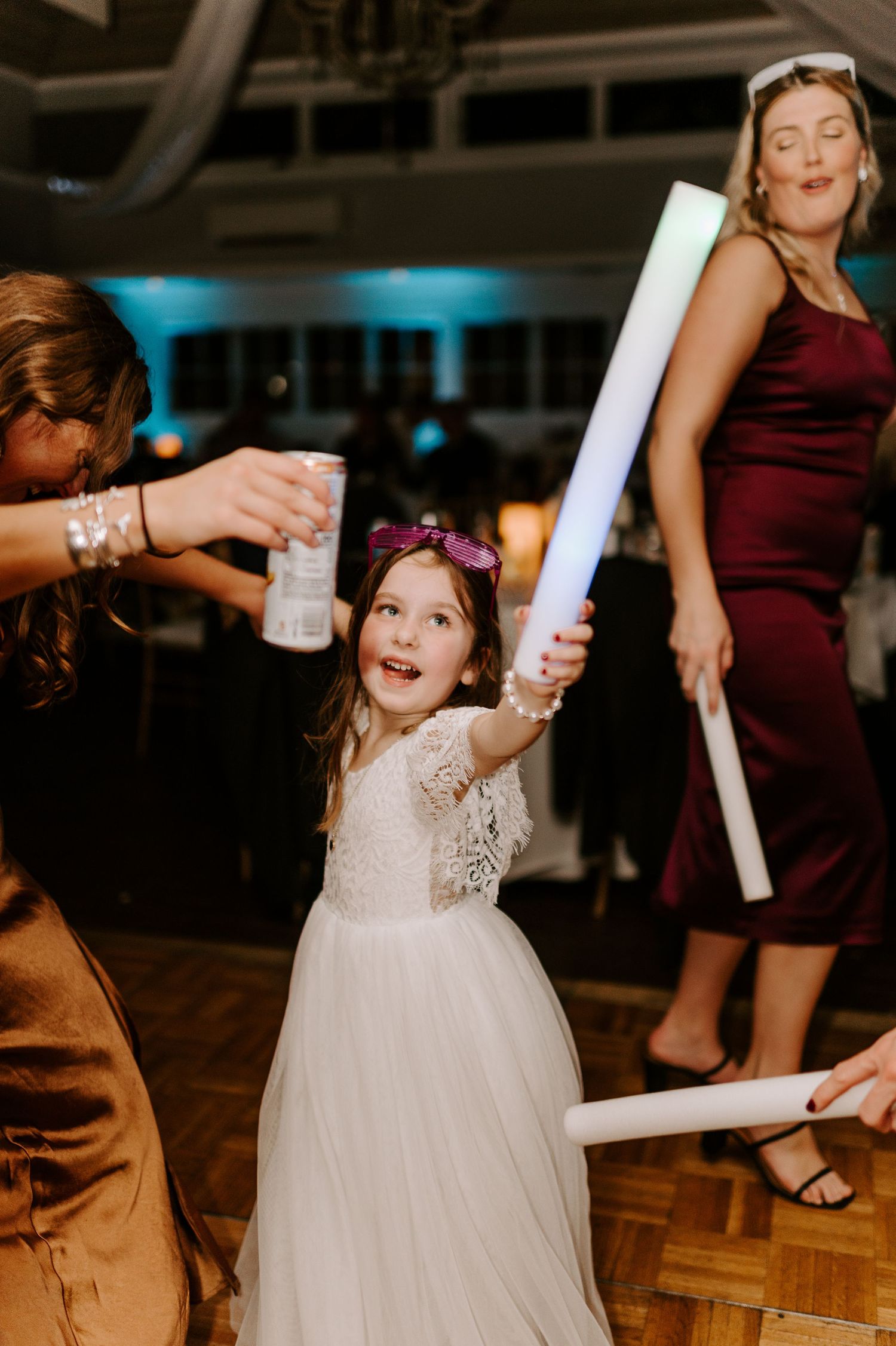 Young flower girl in white dress dancing with light sticks at wedding reception.