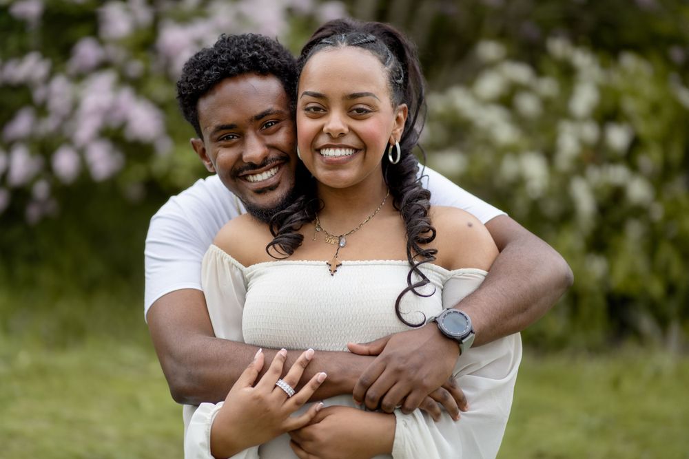 A couple embraces and smiles together outdoors wearing white clothing against a floral background.