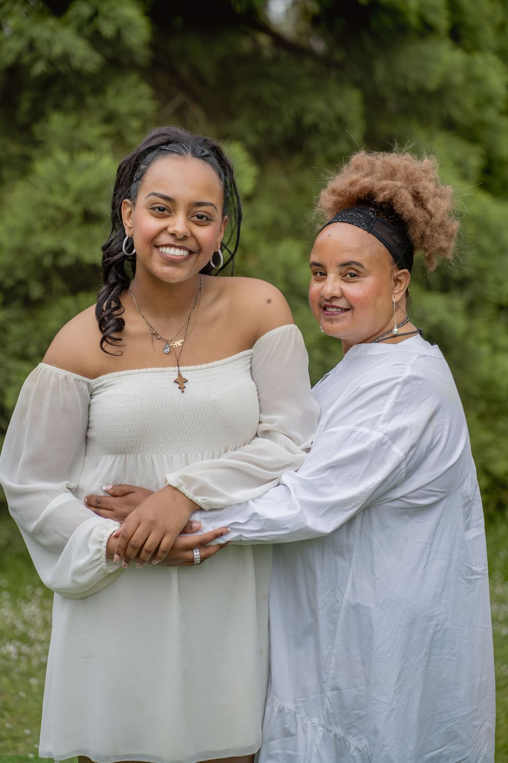 Two women in white dresses embrace while smiling in an outdoor garden setting.