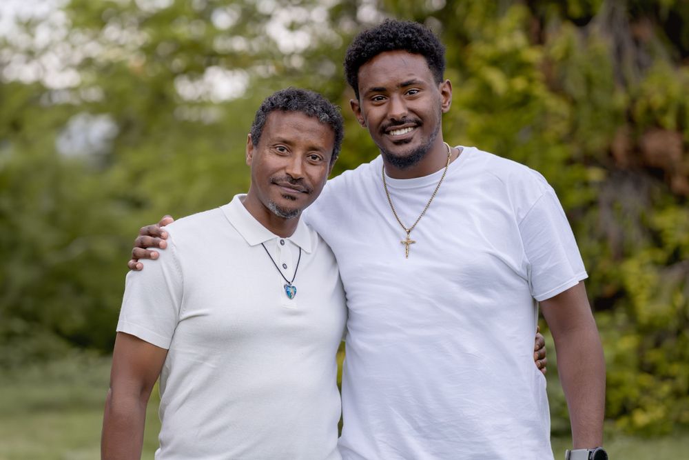 Two people wearing white t-shirts smile together outdoors in a garden setting.