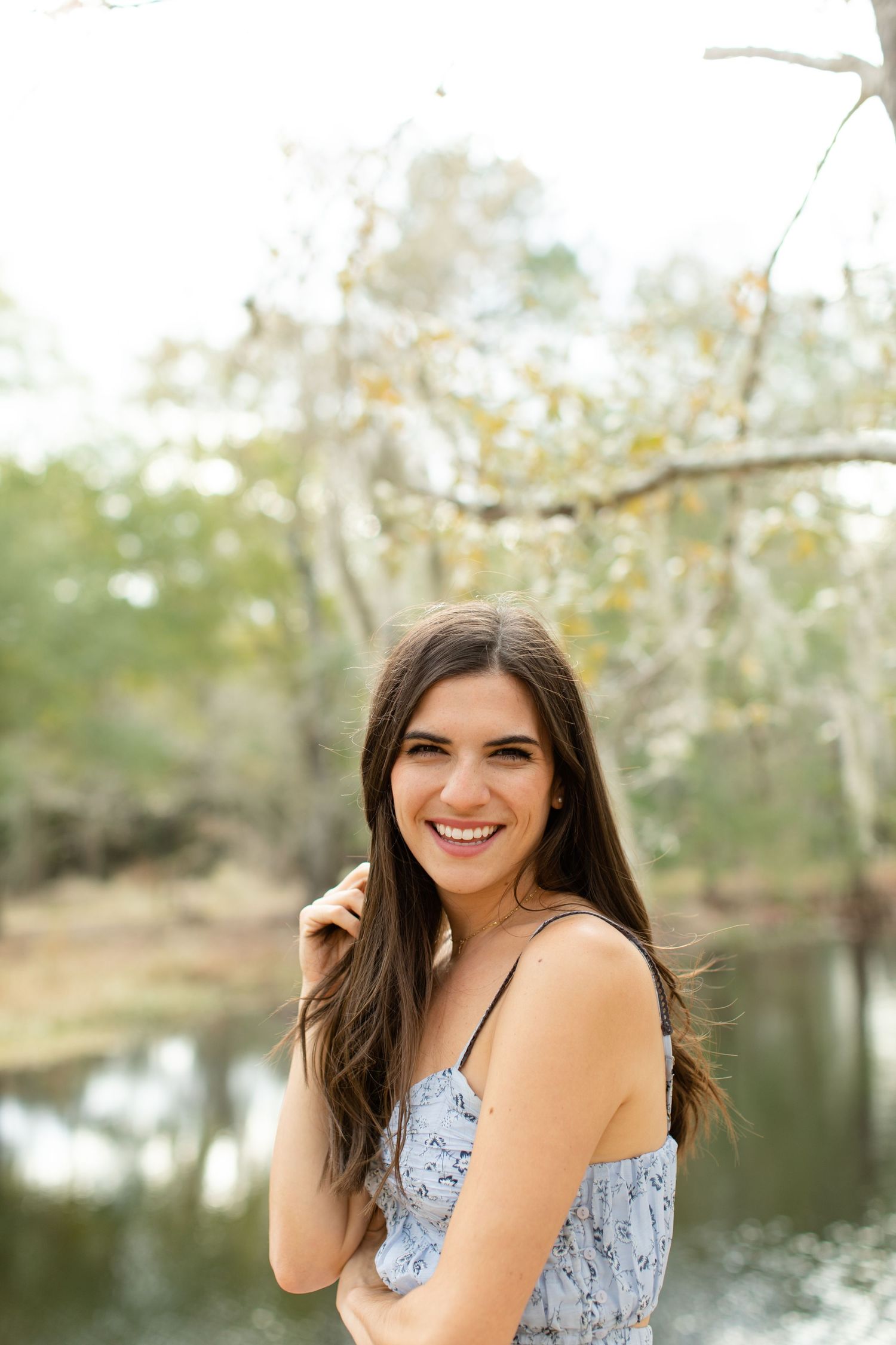 Portrait of woman in blue floral dress laughing outdoors by a lake with natural bokeh background.