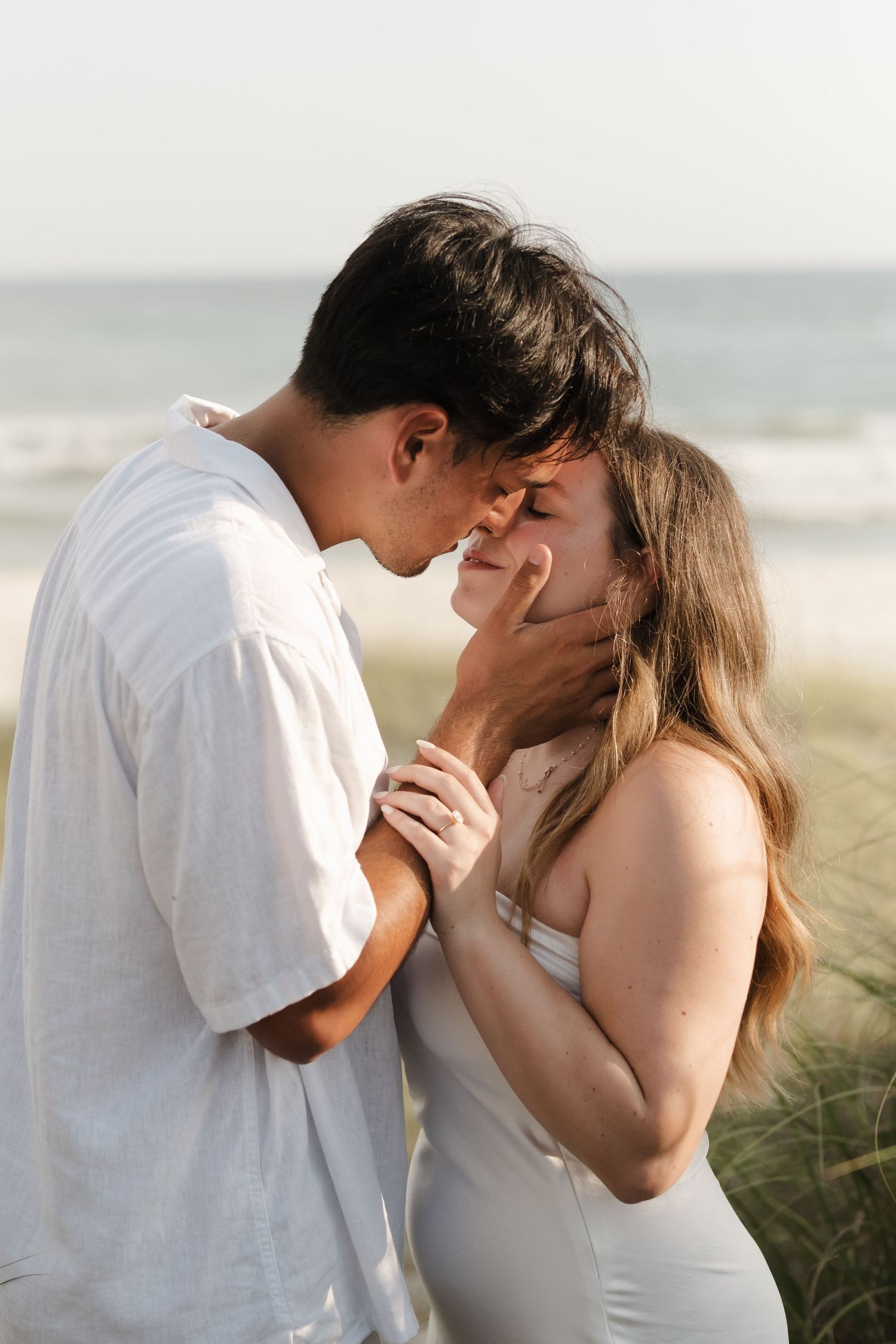 Intimate moment between couple in white attire against coastal background.