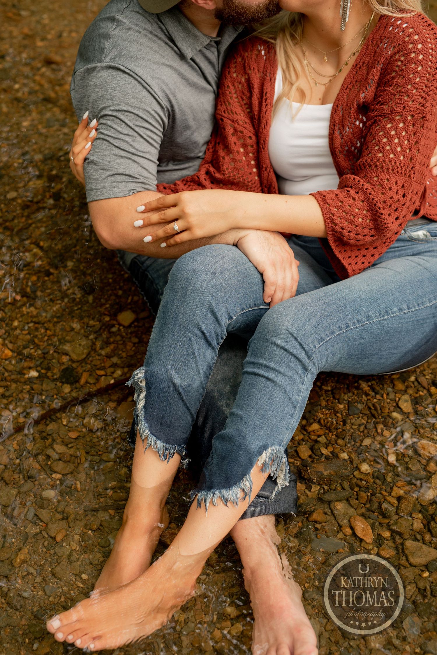 A close-up shows a couple's bare feet and casual attire as they sit together by a creek.