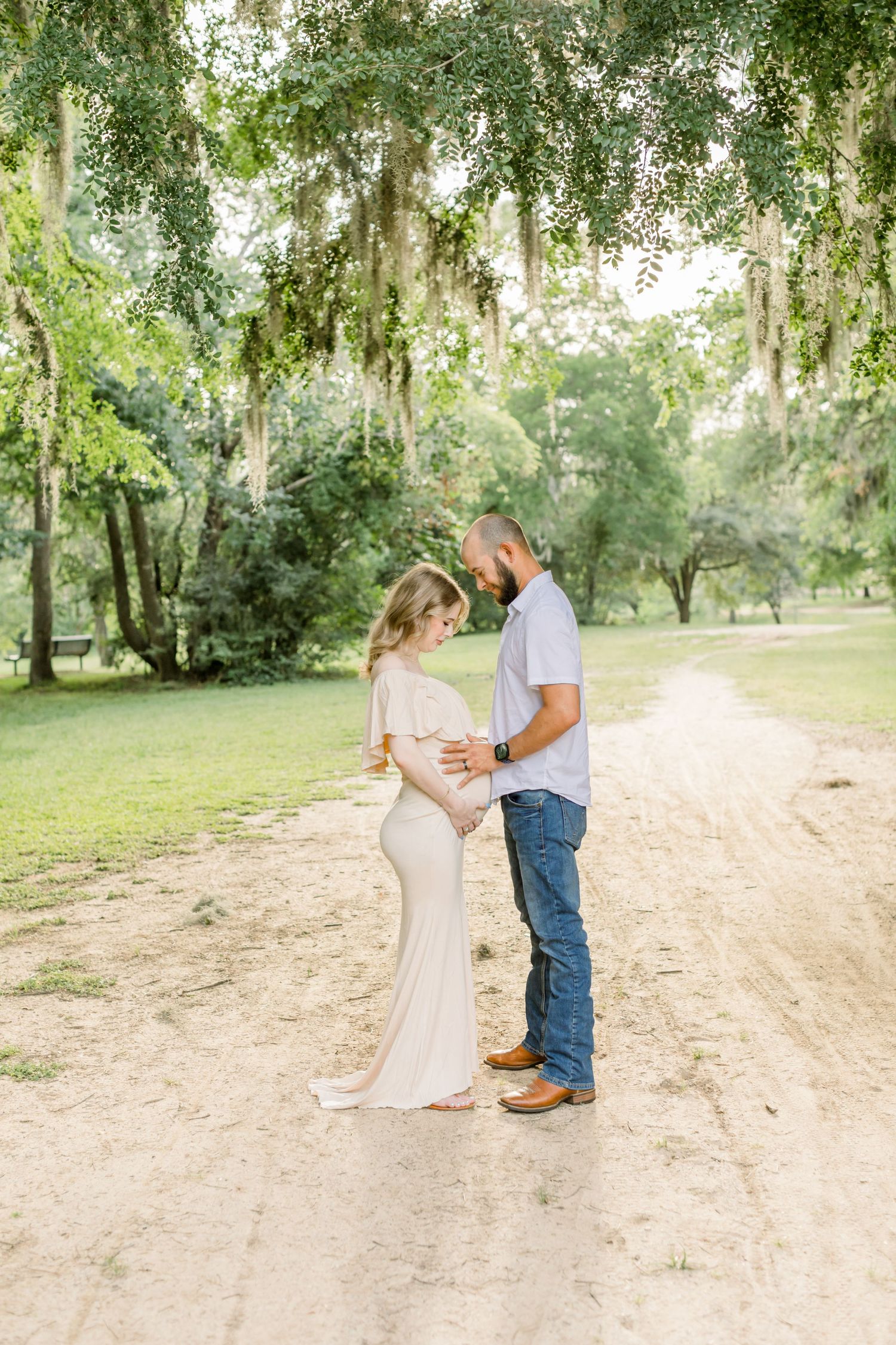 A tender maternity moment captured on a sunlit dirt path beneath moss-covered trees.