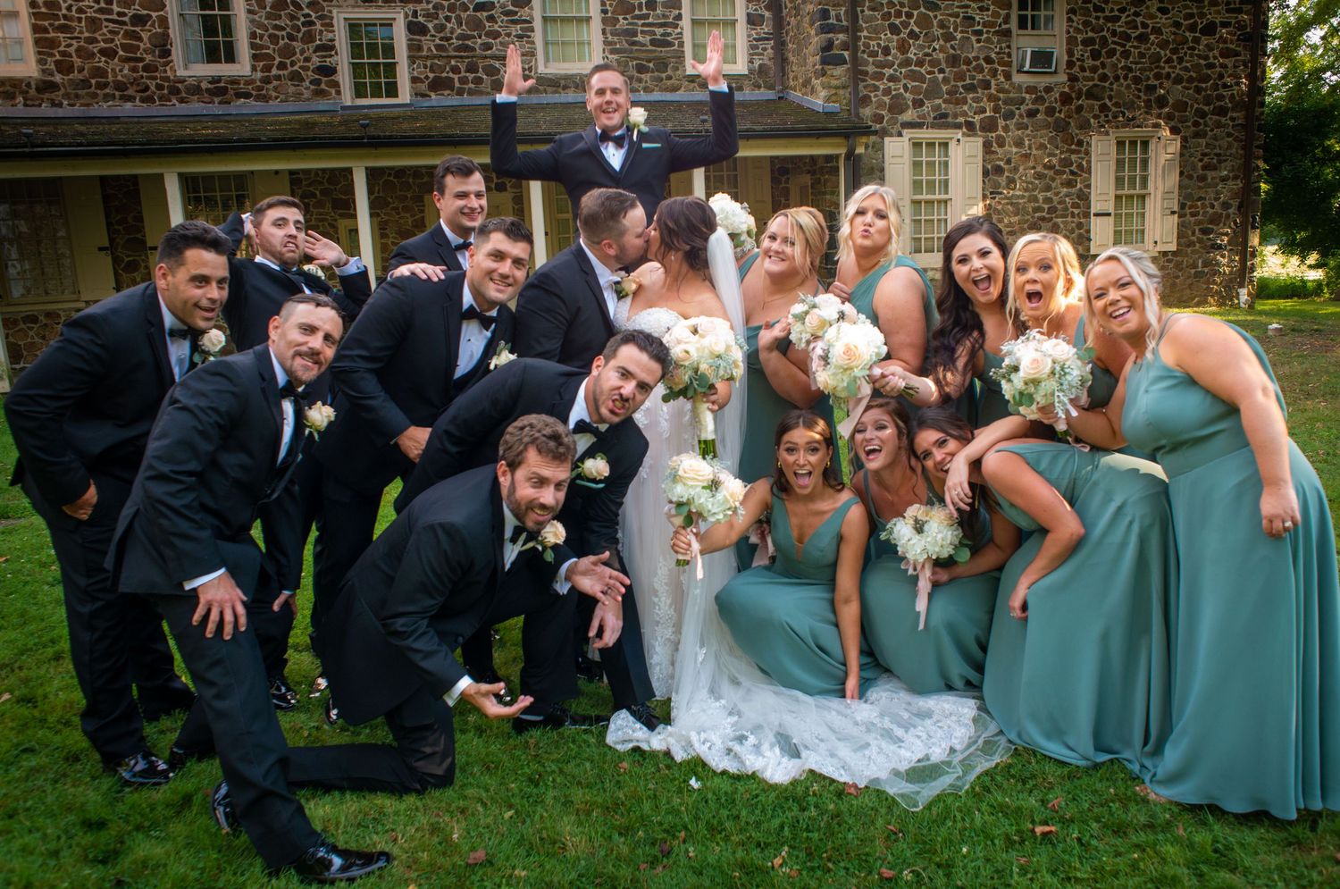 Wedding party in blue dresses and dark suits poses playfully on the lawn of a stone building.