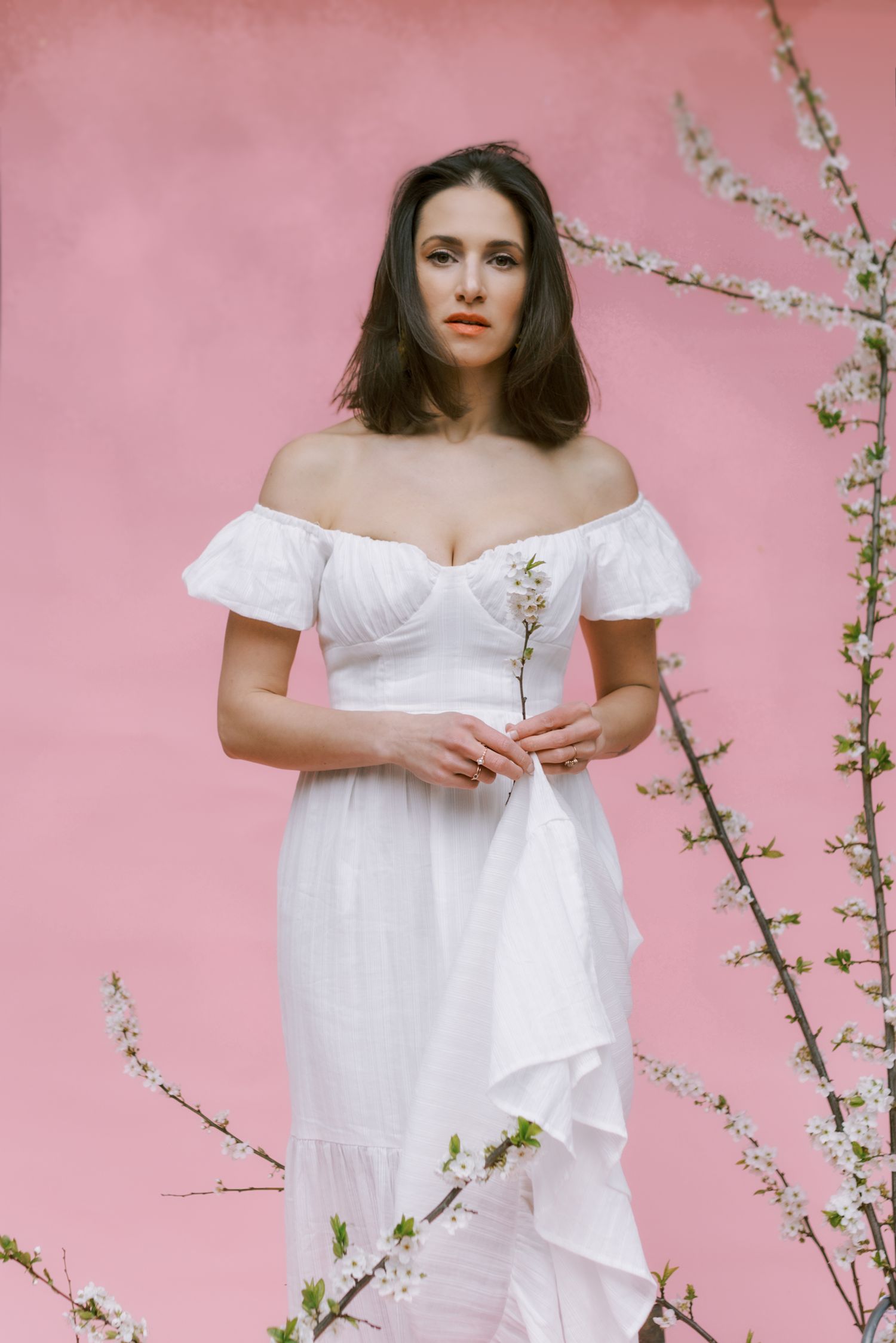 White off-shoulder dress portrait against pink wall with spring cherry blossoms.