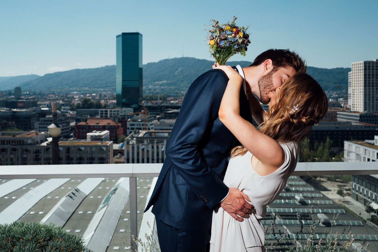 Couple embraces on rooftop with cityscape and mountains in background.