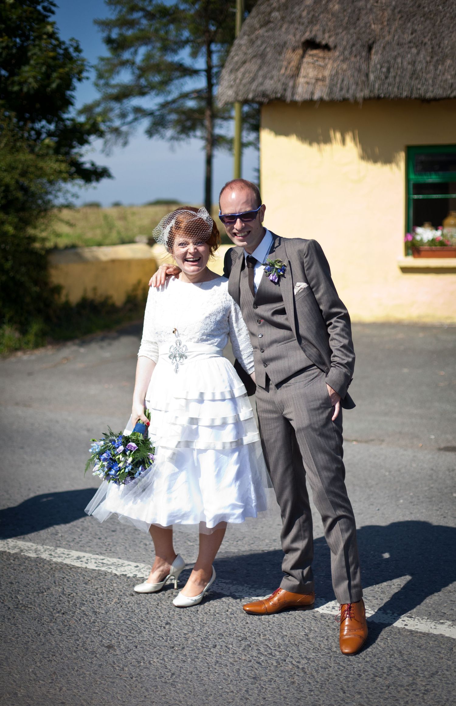 A couple poses outside a rustic thatched-roof cottage on their wedding day.