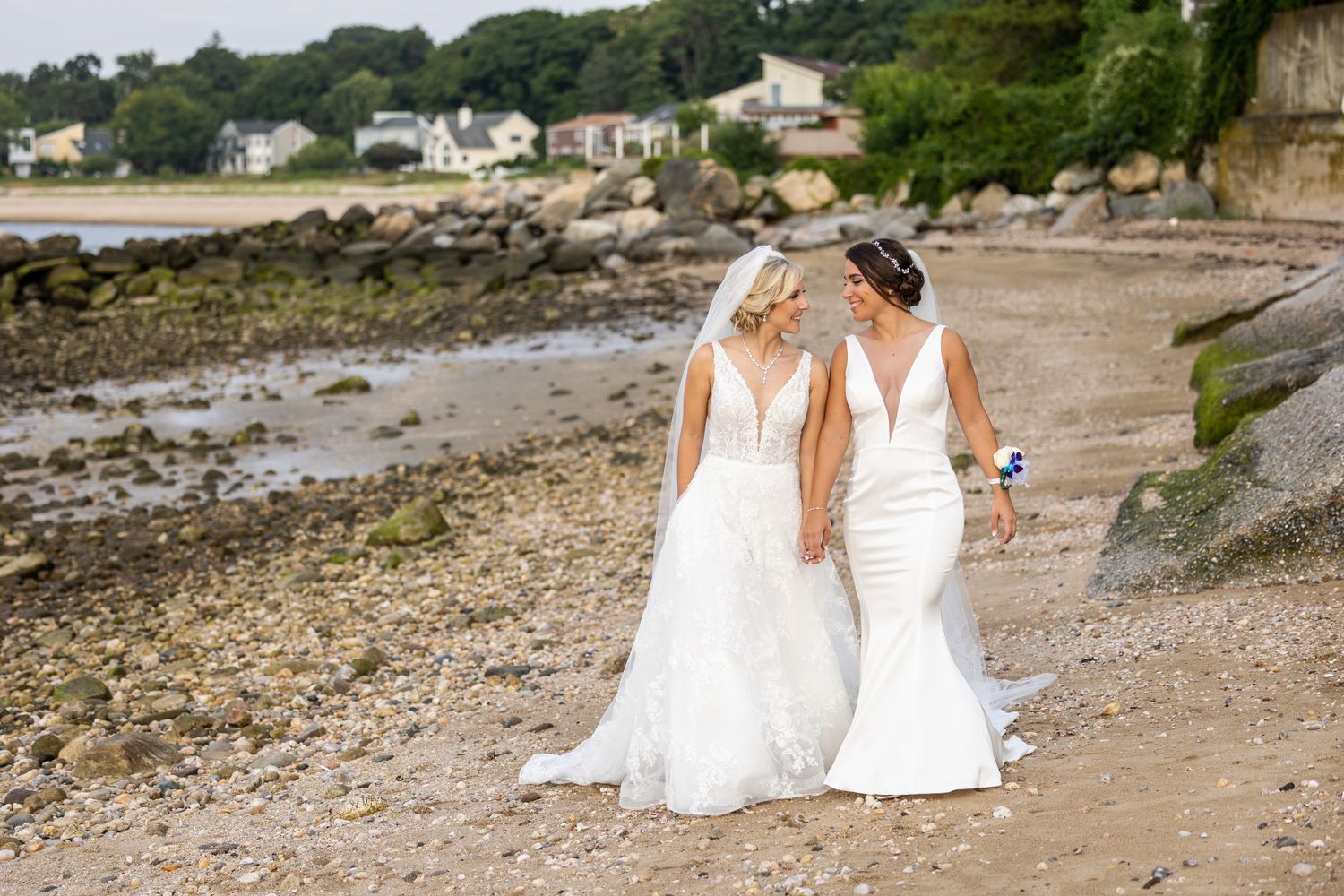 Two brides in white dresses walk along rocky beach shoreline holding hands.