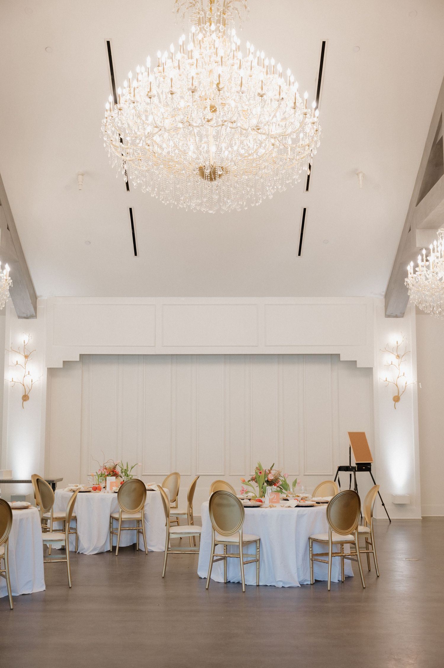Elegant wedding reception venue with crystal chandeliers and round dining tables set against white walls.