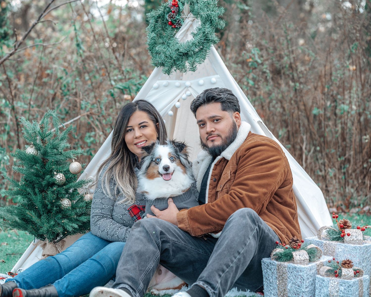 Holiday scene with couple and dog sitting by teepee tent with Christmas decorations and presents.