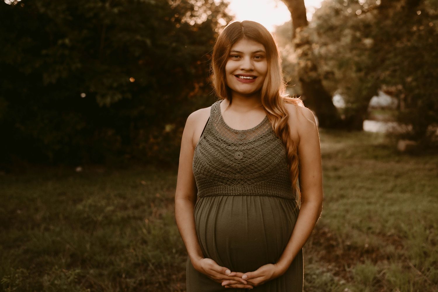 Expectant mother in olive maxi dress poses in golden sunset light.