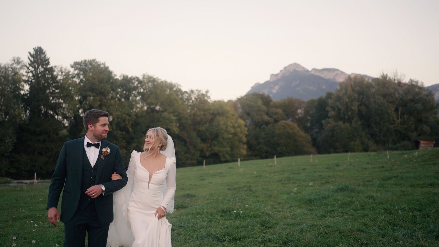 Bride and groom walk together on grassy field with mountain vista in background at sunset.