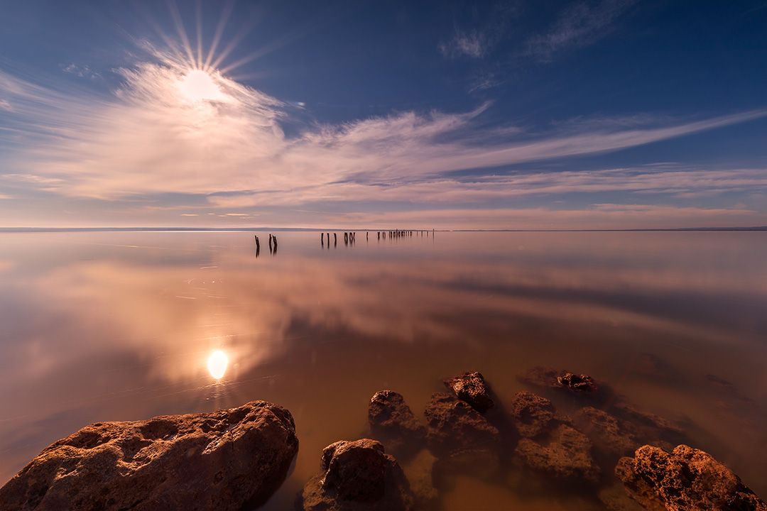 Sunset reflects on calm waters with silhouetted figures and rocks in the foreground creating a mirror-like effect.