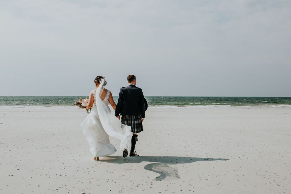 A couple in wedding attire walking along a pristine white sandy beach with ocean waves in the background.