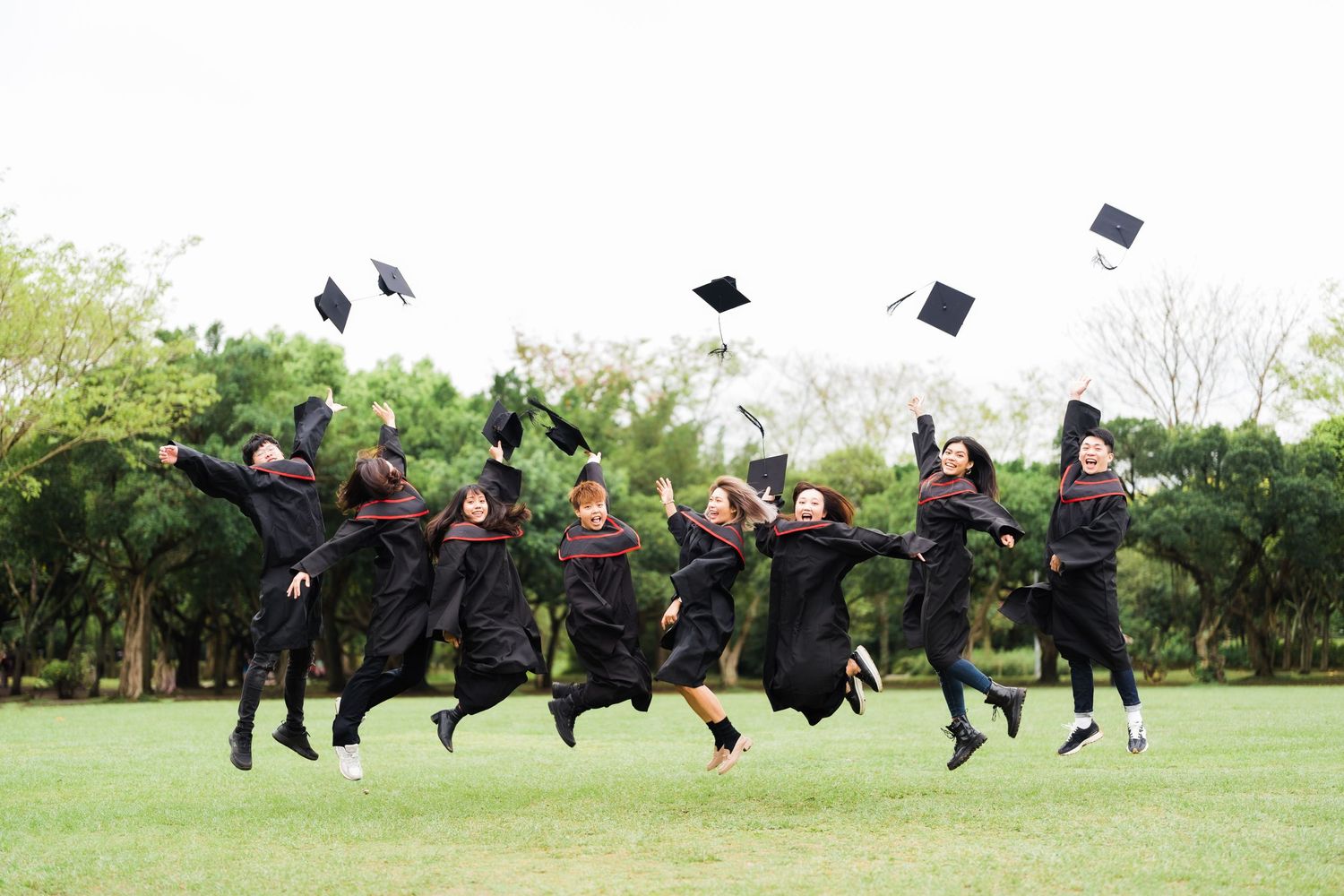 Graduates in black robes jumping with joy while throwing their caps in the air on a green lawn.