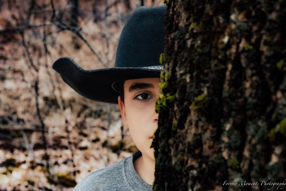 Creative portrait showing half-hidden face behind tree wearing black cowboy hat.