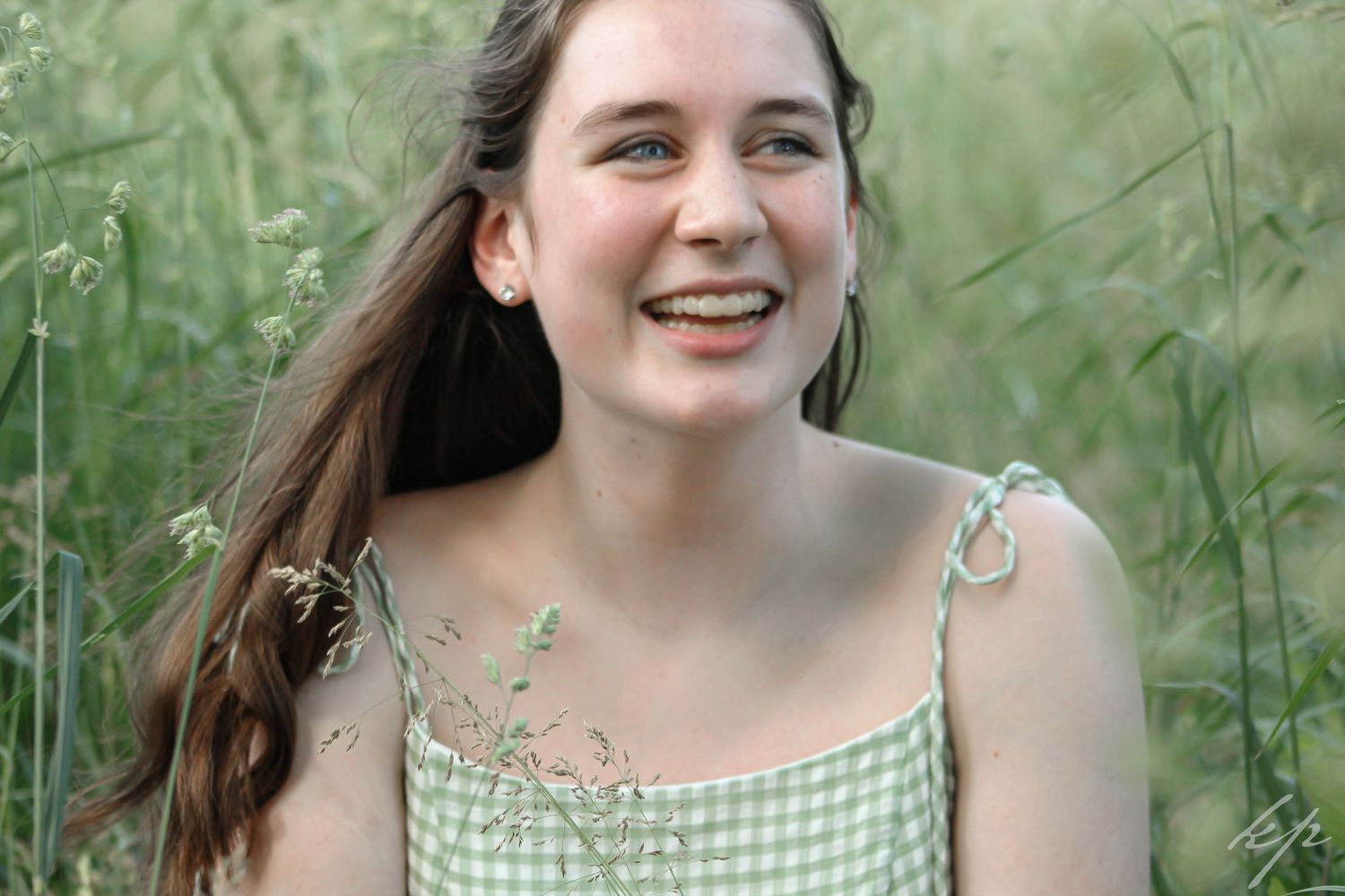 Young woman in a green floral dress poses outdoors against a natural backdrop of tall grass and greenery.