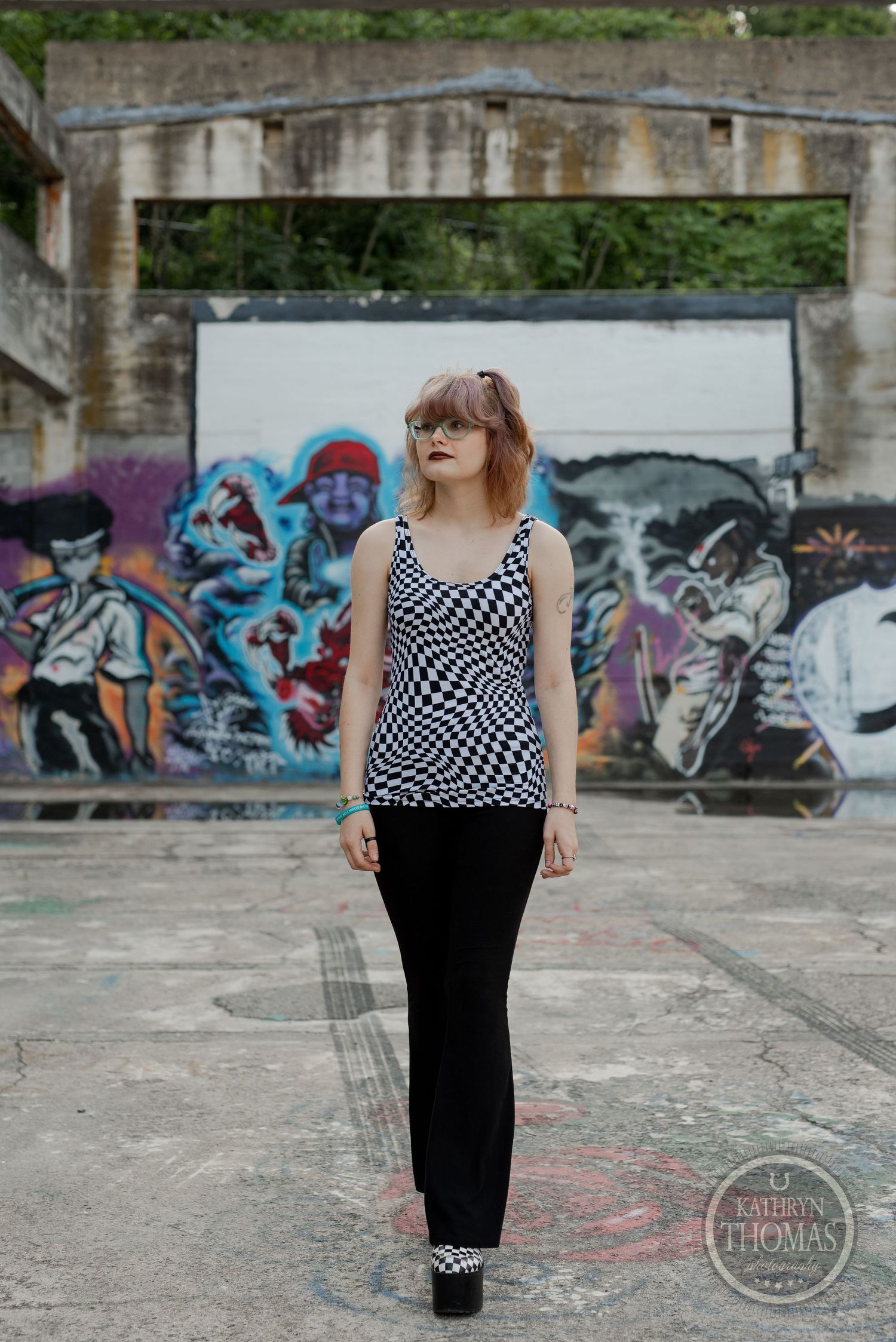 Model in geometric pattern tank top poses in front of urban graffiti wall under concrete architecture.