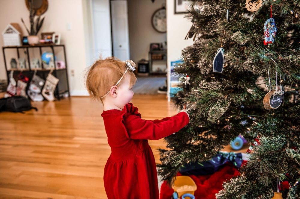 Small child in red dress decorating Christmas tree in living room.