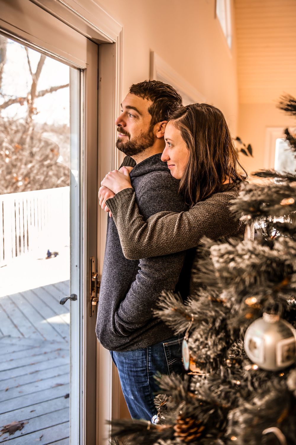 Parent and baby gaze out snowy window together while warmly dressed in winter clothing.