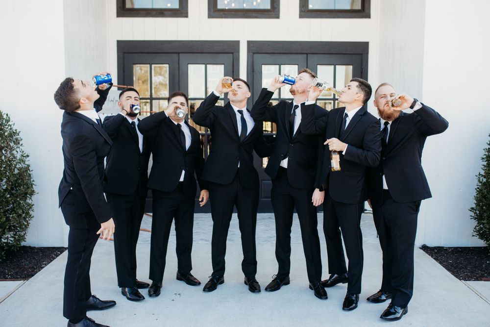 Group of groomsmen in black suits taking celebratory shots outside white wedding venue.