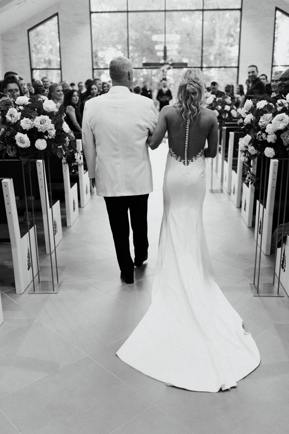 Black and white photo of bride in backless gown walking down church aisle with escort at wedding ceremony.