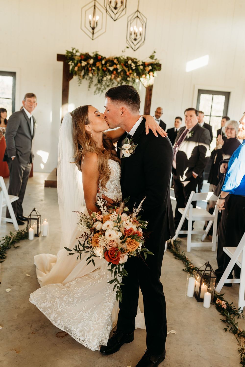 Newlyweds share first kiss in aisle of rustic wedding chapel adorned with floral arch.