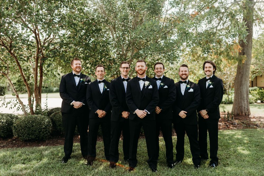 Groom and six groomsmen in black tuxedos with bow ties posing together on green lawn.