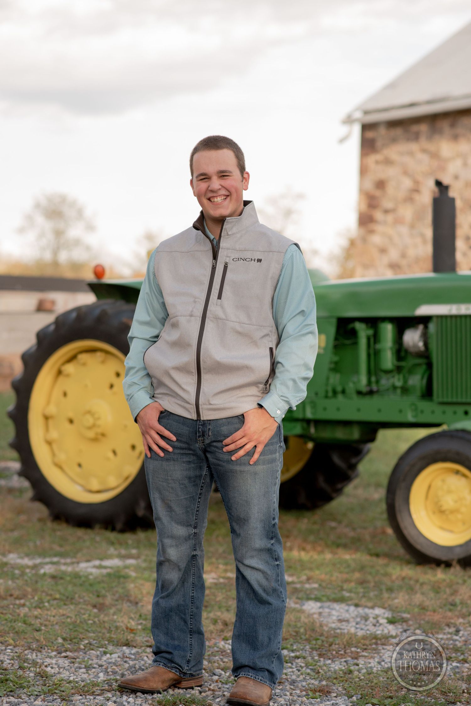 Person in vest and jeans poses casually in front of classic green John Deere tractor.