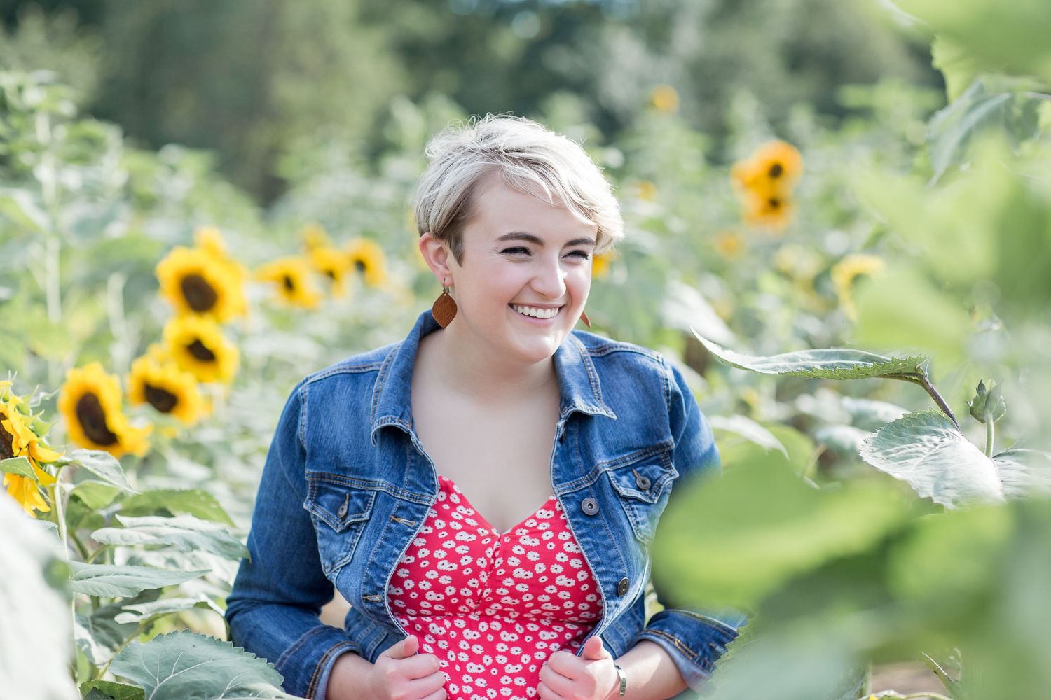 Smiling young woman with short blonde hair in a red floral dress and denim jacket in a sunflower field.