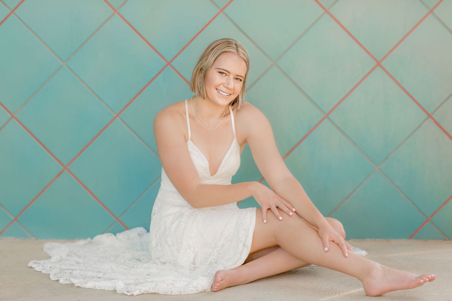 Smiling blonde girl in a white lace mini dress sits barefoot against a teal diamond-patterned wall.