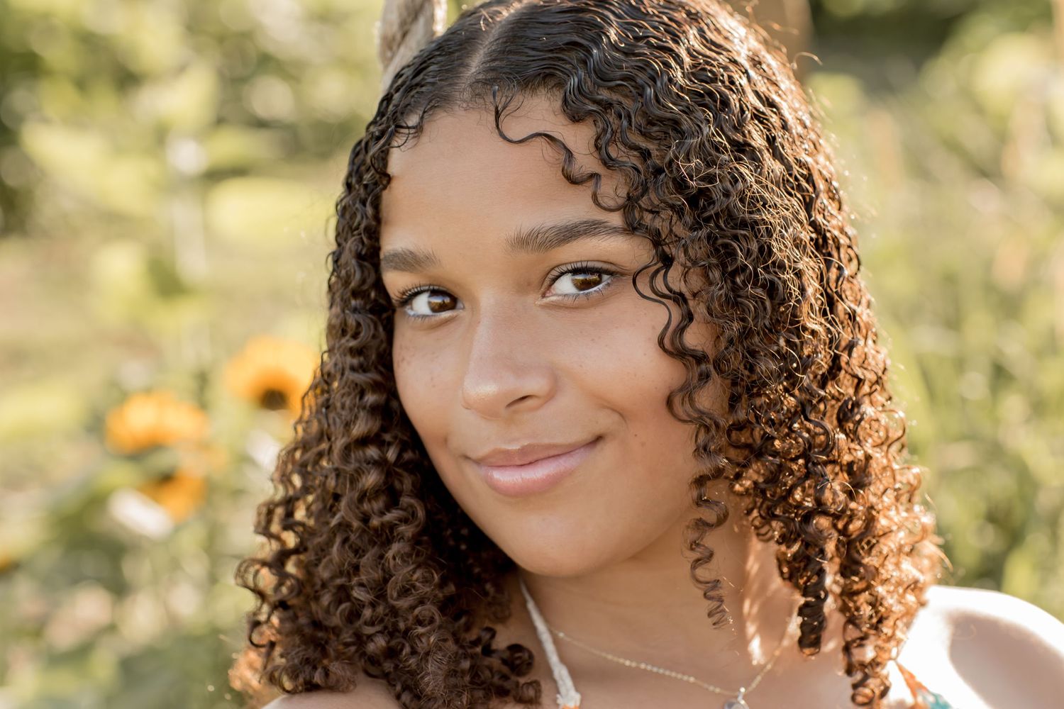 Close-up portrait of a young woman with curly brown hair smiling softly among sunflowers outdoors.