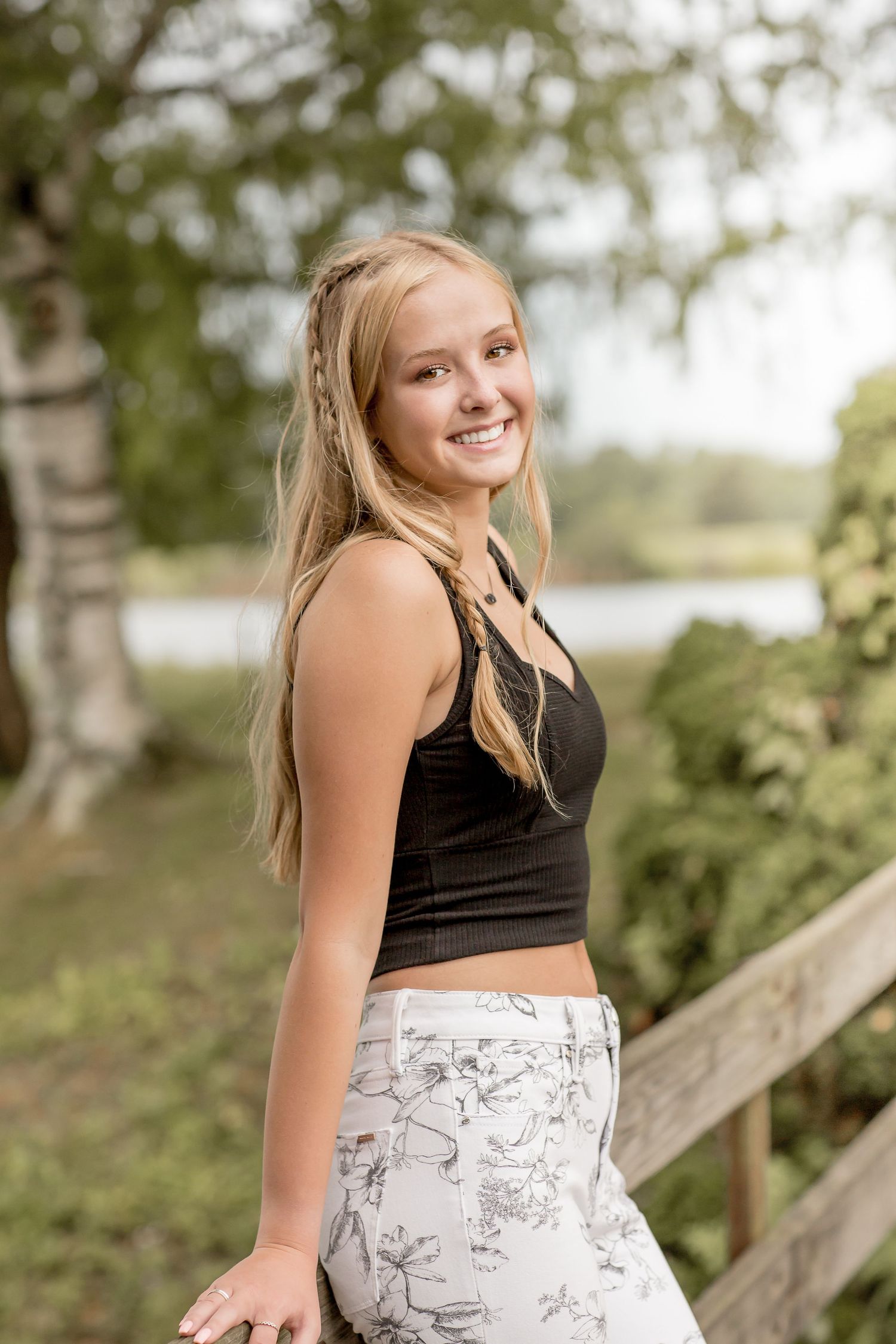 Smiling blonde teen girl in a black crop top and floral pants poses outdoors near a wooden fence.