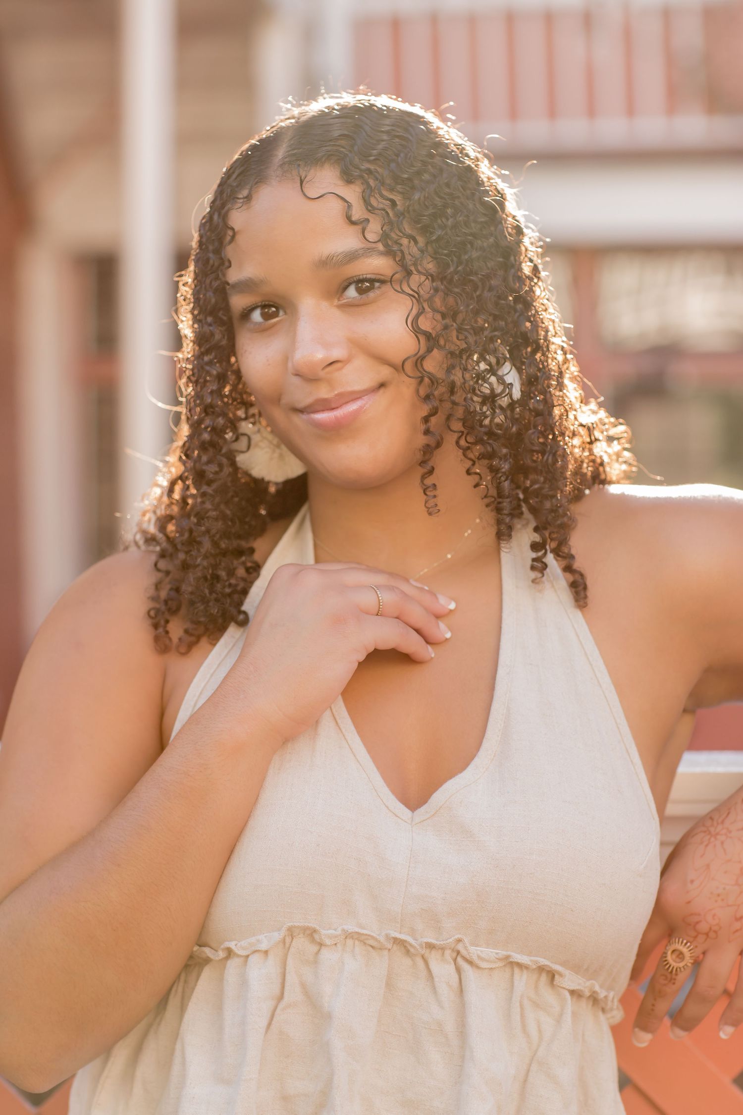 Young woman with curly hair in a beige halter dress poses in warm golden sunlight outdoors.