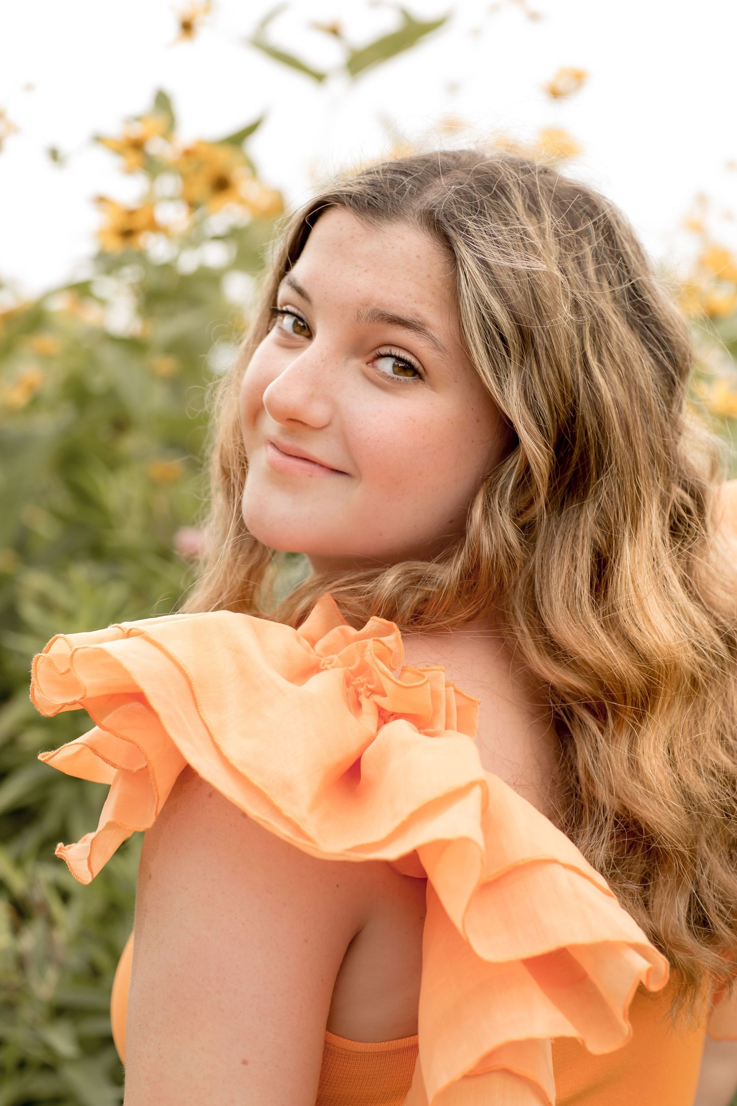 Smiling teen girl with wavy hair wears an orange ruffled off-shoulder dress in a sunflower field.