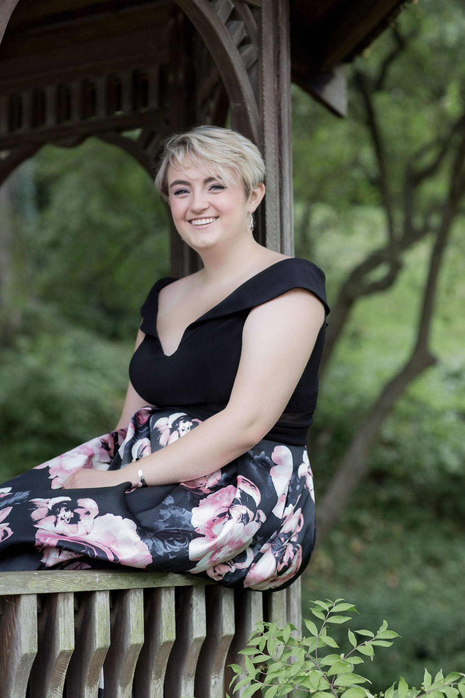 Smiling blonde woman in black top and floral skirt sitting in wooden gazebo surrounded by green trees.