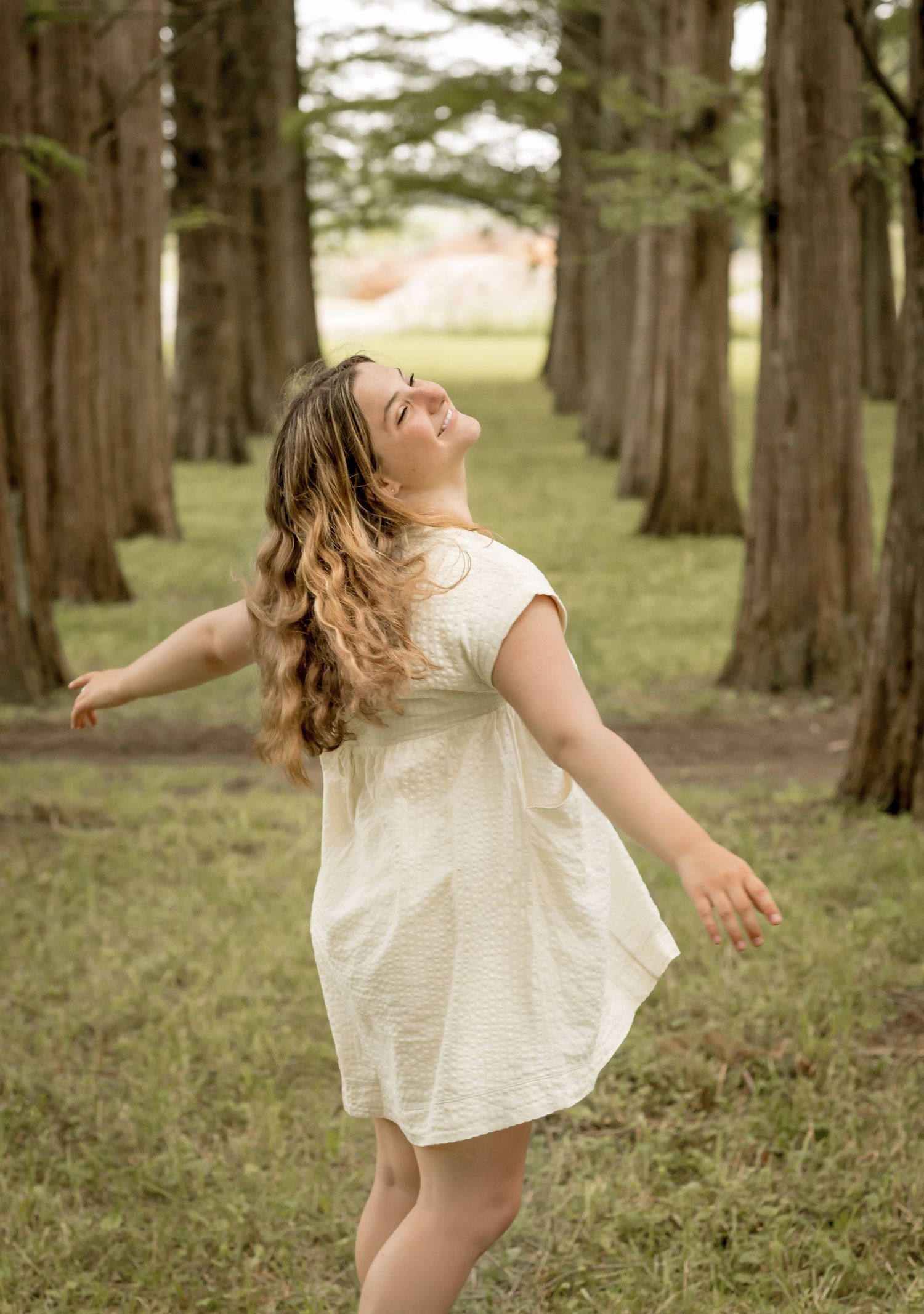 Young woman in white dress twirling joyfully between tall trees in a grassy outdoor setting.