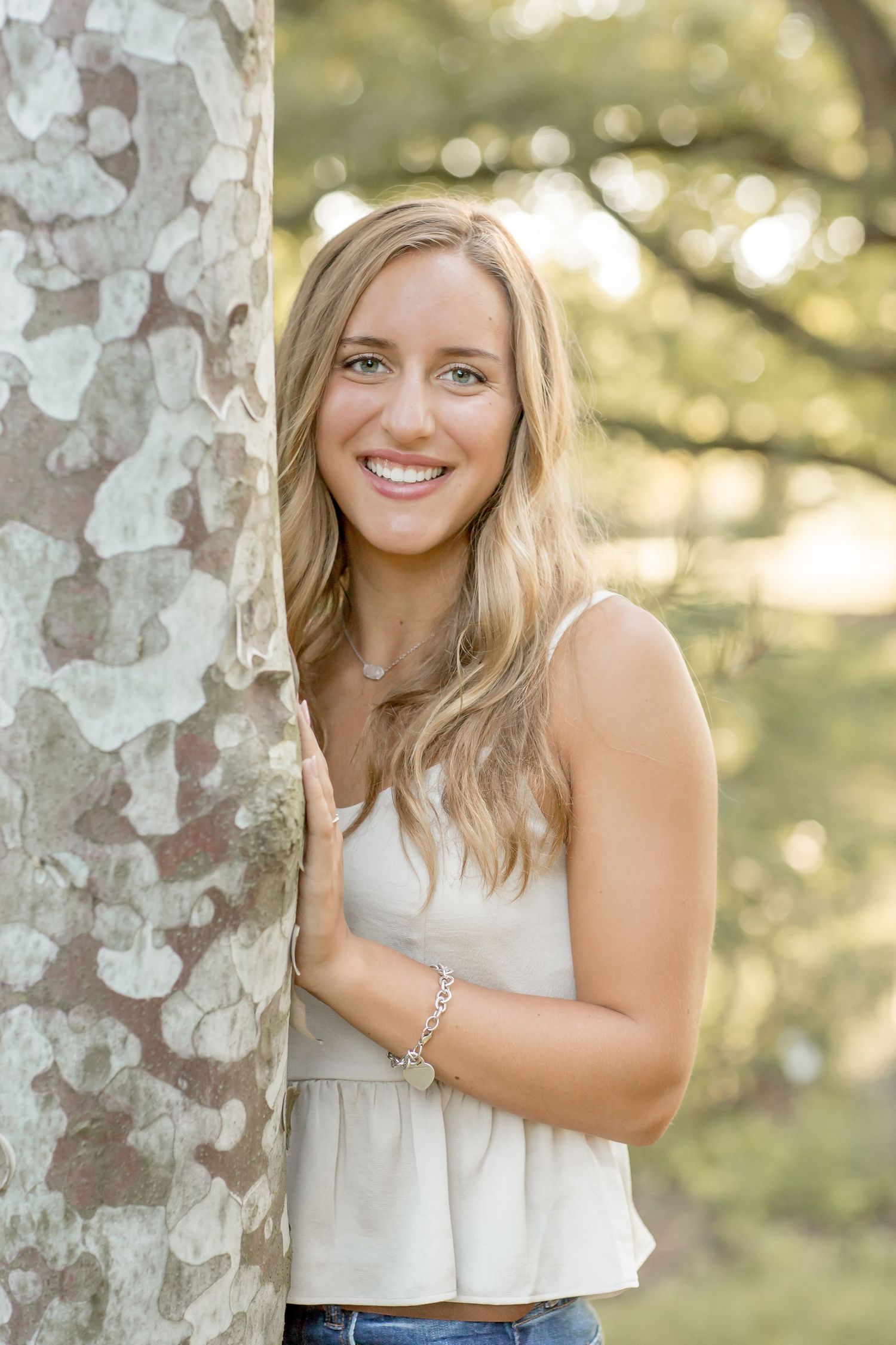 Smiling blonde teen girl in a white tank top leans against a tree trunk in a sunny outdoor setting.