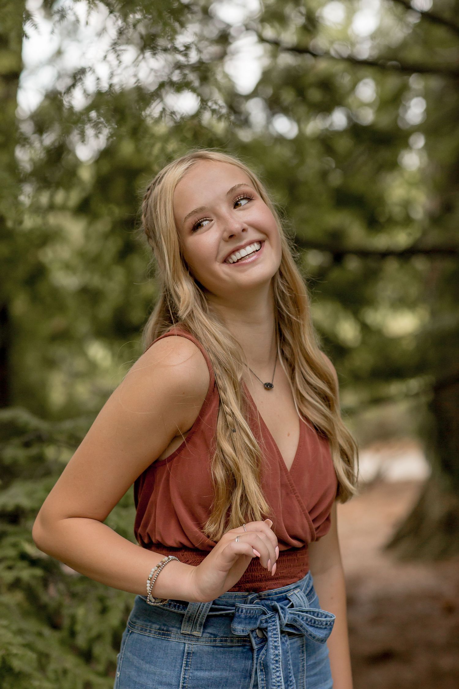 Smiling teenage girl in rust top and denim skirt posing among tall green trees on a forest path.